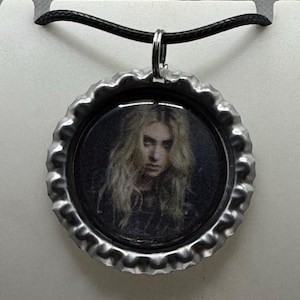 May include: A silver-colored bottle cap pendant necklace featuring a portrait of a person with long blonde hair. The pendant hangs from a black cord necklace. The image is set inside the bottle cap.