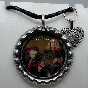 May include: A pendant necklace featuring a circular pendant with a movie scene image, a silver-colored heart charm, and a black cord. The circular pendant is made from a bottle cap. The image shows two people in a scene from a movie.