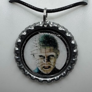 May include: A silver-colored bottle cap pendant necklace featuring a horror movie character image. The pendant hangs from a black cord. The image is of a pale-skinned person with pins in their head and face.