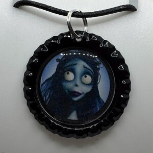 Puede incluir: Colgante de tapón de botella negro con una imagen de dibujos animados de una mujer de piel azul con ojos grandes y cabello oscuro. El colgante cuelga de un collar de cordón negro con una anilla plateada.