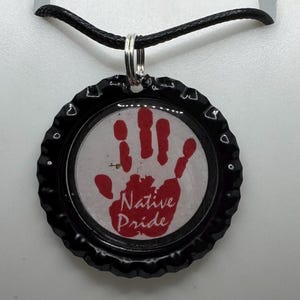 May include: A black bottle cap pendant with a red handprint and the words "Native Pride" on a white background. The pendant hangs from a black cord necklace.