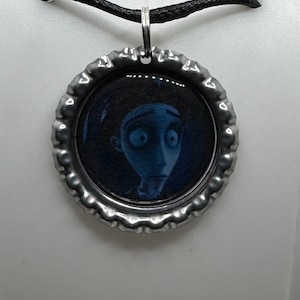Puede incluir: Collar con colgante plateado que presenta un colgante circular con la cara de un personaje de dibujos animados en tonos azules. El colgante está hecho de una chapa de botella y cuelga de un cordón negro.