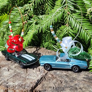May include: Two die-cast toy car ornaments featuring a dark green Mustang with a red bow and a light blue Mustang with a white bow. Each car carries a small bottle brush tree on its roof and includes a wire loop decorated with colorful beads.