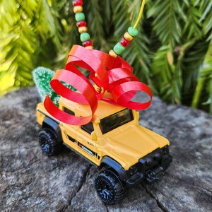 May include: A yellow toy truck with black wheels and accents, adorned with a red ribbon and a small green tree. The truck is suspended by a yellow string with red, green, and brown beads, suggesting a festive ornament.