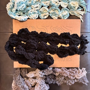 May include: Three crocheted scarves in different colours and textures. The top scarf is blue and white, the middle scarf is black, and the bottom scarf is grey.