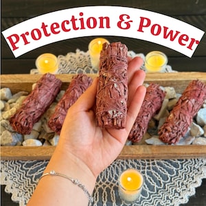 May include: Red sage bundles, wrapped with twine, are displayed in a wooden tray with rocks. The text "Protection & Power" arches across the top. Lit candles add a warm glow to the scene, suggesting a spiritual or wellness theme.