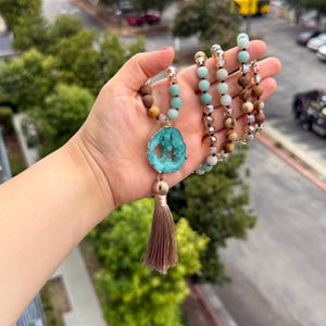 May include: A long beaded necklace featuring a central turquoise-colored geode pendant with a gold-tone rim, accented by a light brown tassel. The strand consists of alternating light blue, brown, and clear faceted beads on a dark cord.