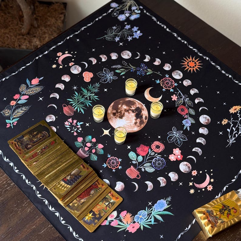 May include: A black tarot cloth featuring a moon phase design with floral and celestial elements. The cloth has a white border and is adorned with a full moon, crescent moons, and various flowers. A deck of tarot cards and candles are arranged on the cloth.