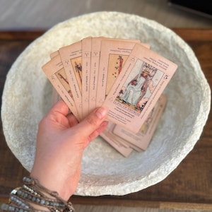 May include: A hand holds a fan of tarot cards, featuring illustrations and text. The cards are in shades of beige and brown, with the "King of Swords" card prominently displayed. The cards are set against a textured, off-white bowl.