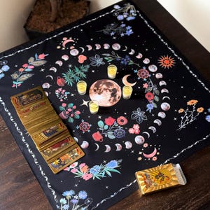 May include: A black tarot cloth featuring a moon phase design with floral and celestial elements. The cloth has a white border and is adorned with colorful flowers, moons, and stars. A deck of tarot cards and a gold-wrapped item are also present.