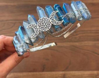 May include: A headband featuring a row of iridescent blue crystal points wrapped in silver wire. A silver-toned metal charm depicting a triple moon symbol with intricate filigree patterns is attached to the center, accented by two delicate silver chains.