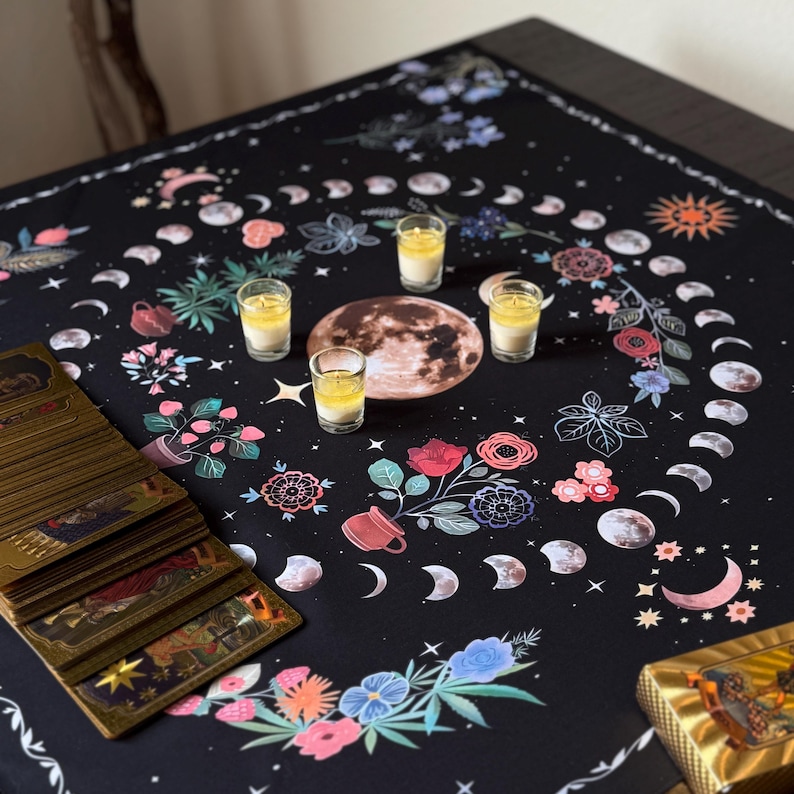 May include: A black tarot cloth featuring a moon phase design with floral and celestial elements. The cloth has a central moon graphic, surrounded by flowers, moons, and stars. Several shot glasses with yellow liquid are placed on the cloth, along with a deck of tarot cards.