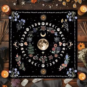 May include: Black square tapestry featuring a detailed moon phase design. The central image is a full moon surrounded by crescent moons, stars, and floral elements in shades of white, pink, and blue. The tapestry is bordered by a white decorative trim.