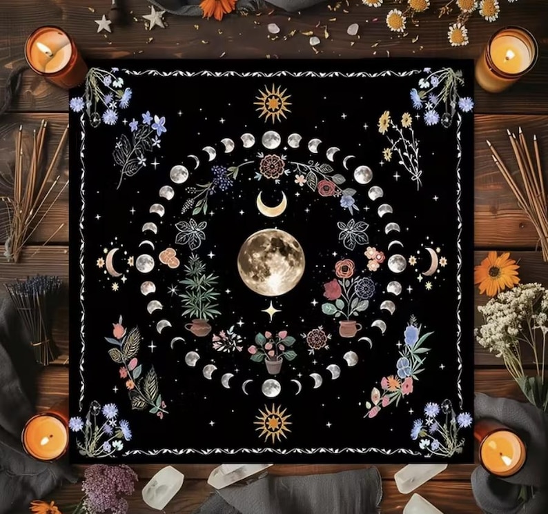 May include: Black square tapestry featuring a detailed moon phase design. The central image is a full moon surrounded by crescent moons, stars, and floral elements in shades of white, pink, and blue. The tapestry is bordered by a white decorative trim.