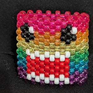 May include: A colorful beaded bracelet featuring a cartoon face design. The bracelet is made of small, round beads in a rainbow of colors, including pink, orange, yellow, green, blue, and purple. The face has black eyes, white teeth, and a red mouth.