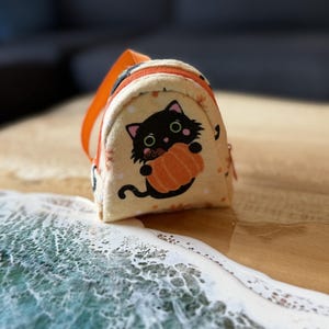 May include: A small, cream-colored fabric backpack featuring a graphic of a black cat holding an orange pumpkin. The bag has an orange zipper, orange straps, and a pattern of small orange leaves and white dots across the light-colored fabric.