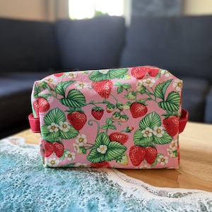 May include: A rectangular fabric pouch featuring a repeating pattern of red strawberries, green leaves, and white strawberry blossoms on a light pink background. The pouch has red fabric side tabs and a structured shape with visible seams.