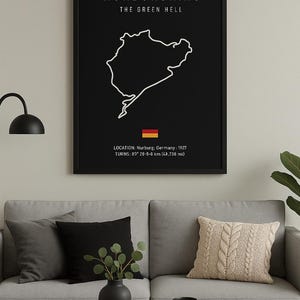 Puede incluir: Póster enmarcado en negro con el contorno blanco del circuito de carreras de Nürburgring. El texto dice "Nürburgring, The Green Hell". Incluye ubicación, curvas y distancia en km (42.738 mi). La bandera alemana está en la parte inferior.