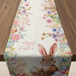 May include: Easter-themed table runner featuring a bunny, Easter eggs, and floral designs in pastel colors. The words "Happy Easter" are printed on the runner. The runner is made of a soft fabric and is designed to decorate a table.