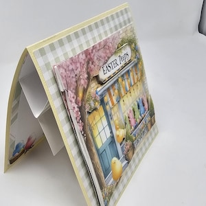 May include: A handmade card with a pop-up design featuring a small shop with a sign that reads "Easter Peeps". The shop is decorated with pastel-colored eggs and flowers. The card is made with a checkered pattern paper and has a yellow border.