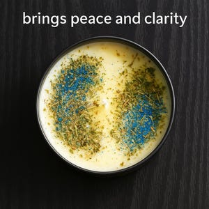 Op de afbeelding: Een ronde kaars in een zwarte container met een crème-kleurige wasbasis. De kaars is versierd met groene en blauwe gedroogde kruiden en de tekst "The Peaceful 1 brings peace and clarity".
