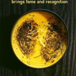 May include: A round, yellow candle in a black container, adorned with dried herbs. The text "THE STAR brings fame and recognition" is displayed in yellow above the candle. The candle sits on a dark, textured surface.