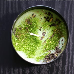 May include: A round, green candle in a black metal container. The candle is decorated with green glitter and dried herbs. The text "THE ABUNDANT 1 attracts money, luck and abundance" is printed above the candle.