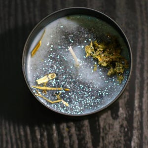 Op de afbeelding: Een ronde, zwarte kaarsencontainer gevuld met een lichtblauwe wasbasis, glitter en gedroogde kruiden. Een witte lont is gecentreerd in de was. De kaars heeft een rustieke, natuurlijke uitstraling.