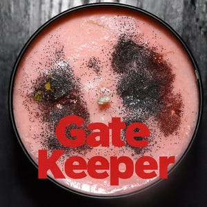 Op de afbeelding: Een ronde, roze kaars in een zwarte tin. De kaars is versierd met zwarte glitter en gedroogde kruiden. De tekst "Gate Keeper" is in het rood op de bodem van de kaars gedrukt.