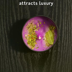 Op de afbeelding: Een ronde, paarse kaars met gouden glitter, kruiden en een laurierblad. De kaars zit in een zwarte metalen houder. De tekst "THE LUXURY 1 attracts luxury" staat bovenaan.