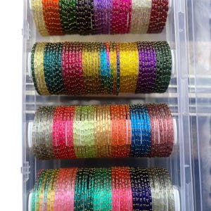Puede incluir: Un recipiente de plástico transparente lleno de cuatro filas de pulseras de vidrio coloridas. Cada fila presenta una variedad de colores, incluyendo rojo, naranja, amarillo, verde, azul y morado. Las pulseras están adornadas con pequeñas cuentas doradas.