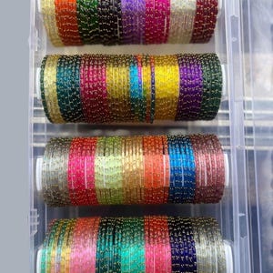 Op de afbeelding: Een verzameling veelkleurige glazen armbanden gerangschikt in vier horizontale rijen in een doorzichtige plastic opbergdoos. Elke armband heeft een getextureerd oppervlak met kleine goudkleurige accenten en toont een verscheidenheid aan levendige tinten, waaronder rood, groen, paars, geel, blauw, roze en helder glas.