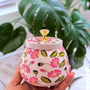 Op de afbeelding: Een kleine, ronde witte keramische pot met een herhalend patroon van roze lotusbloemen en groene waterleliebladeren. Het deksel is bekroond met een doorzichtige, gefacetteerde, diamantvormige knop op een goudkleurige metalen voet.