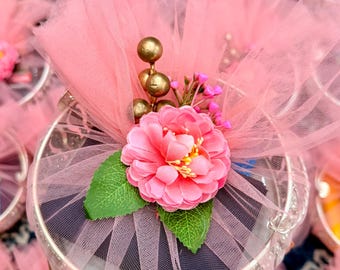 May include: Pink tulle-wrapped favors, each adorned with a large pink flower, green leaves, and gold accents. The favors are presented in round, silver-rimmed containers, creating a delicate and festive appearance, perfect for celebrations.