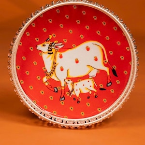 May include: A decorative round tray with a red background featuring a white cow and calf illustration. The tray has a silver beaded rim and small paisley patterns. The cow has red handprints on its body.