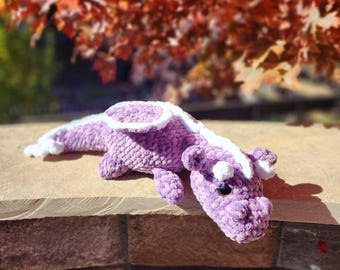 Puede incluir: Un juguete de dragón de peluche hecho a mano en tonos morados y blancos. El dragón está acostado sobre una superficie de piedra, con sus alas y cabeza visibles. El fondo presenta follaje otoñal borroso.