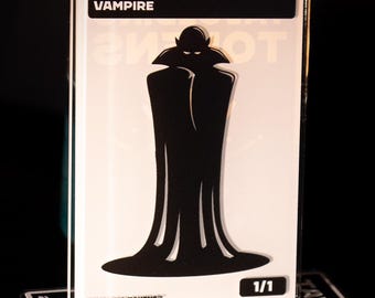 May include: A white and black Vampire token card with a silhouette of a vampire in a long coat. The card is labeled "VAMPIRE" at the top and "1/1" in the corner. The card is on top of a black and white coaster with the text "TAVOLDING TOKENS".