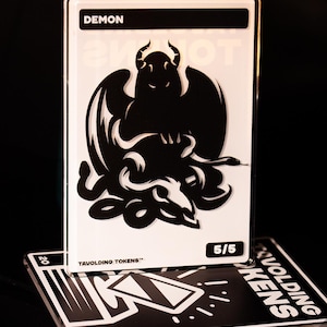 May include: A white card with the word "DEMON" at the top and a black illustration of a demon with horns and wings. The card is labeled "5/5" and "TAVOLDING TOKENS".