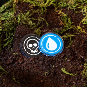 May include: Two bottle caps with graphic designs rest on a bed of soil and moss. One cap features a white skull on a black background, the other a white water droplet on a blue background. The caps have a metallic sheen.