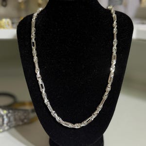 May include: A silver-colored chain necklace with a unique link design is displayed on a black velvet jewelry bust. The necklace has a repeating pattern of rectangular and oval links, creating a textured appearance. The jewelry is showcased against a neutral background.