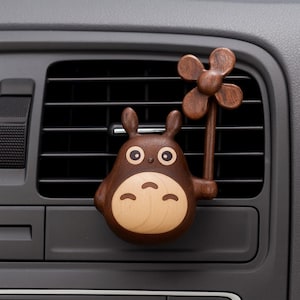 Könnte beinhalten: Ein hölzerner Auto-Lüftungsclip mit einer Cartoonfigur mit großen Augen und einer Blume. Der Clip besteht aus dunkelbraunem und hellbraunem Holz und ist an einer Autolüftung befestigt.