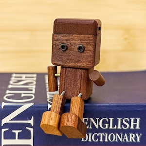 May include: A wooden robot figurine with a dark brown head and body, and lighter brown limbs. The robot has two small, black eye details and is sitting on a blue book with the words "ENGLISH DICTIONARY" on the cover.