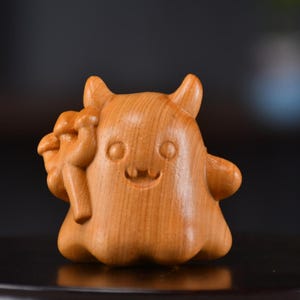 Puede incluir: Pequeña figura de madera de una criatura de dibujos animados con una cara sonriente, pequeños cuernos y sosteniendo un grupo de objetos. La figura está tallada en madera de color claro, con vetas visibles. La criatura tiene un cuerpo redondeado y rasgos sencillos.