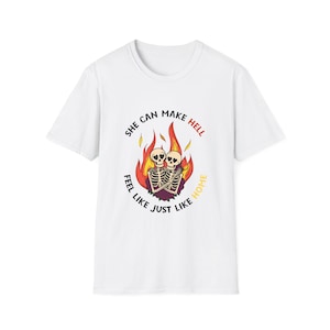 May include: White t-shirt with a graphic of two skeletons in flames. The text reads "She Can Make Hell Feel Like Just Like Home" in a circular design. The design uses red, yellow, and black colours.