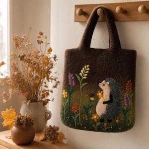 Puede incluir: Un bolso de fieltro marrón oscuro con un bordado que muestra un erizo sosteniendo una flor amarilla en un campo de flores silvestres coloridas y una pequeña mariquita. El bolso tiene dos asas integradas y una forma rectangular con esquinas redondeadas.