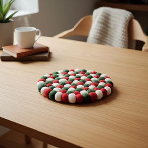 May include: A round trivet made of felt balls in red, green, and white. The trivet is on a wooden table, with a cup of coffee and a plant in the background. The trivet is approximately 8 inches in diameter.