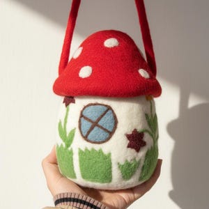 May include: A felt mushroom-shaped bag with a red top and white spots. The bag has a white body with a blue window, green grass, and floral details. It has a red strap for carrying.