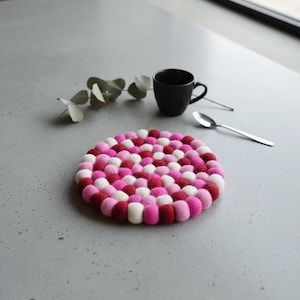 Puede incluir: Un salvamanteles redondo de fieltro compuesto por pequeñas bolas de colores en tonos rosa, rojo y blanco. Una taza negra y una cuchara plateada están al lado. El salvamanteles tiene unos 20 cm de diámetro.