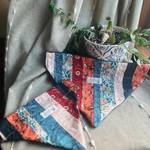 May include: Two colorful dog bandanas with a patchwork design. The bandanas feature a mix of floral, geometric, and animal prints in shades of blue, red, orange, and black. Each bandana has a small white label with text.
