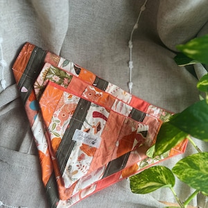May include: Three orange, white, and brown patchwork dog bandanas. The bandanas have a variety of floral and geometric patterns. The bandanas are folded and stacked on top of each other. The bandanas have a label that says "Patchwork & Pups Co."